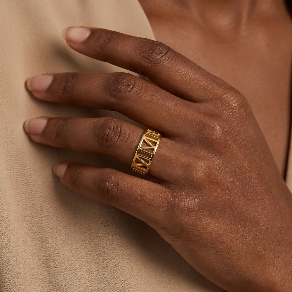 Hand resting on a beige top, wearing a wide gold ring with cut-out Roman numerals