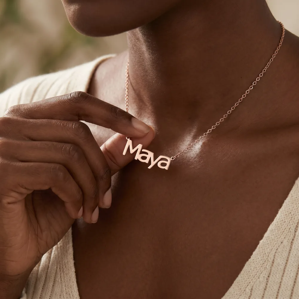 Woman in a cream knit top holding a rose gold block-letter name necklace that reads Maya