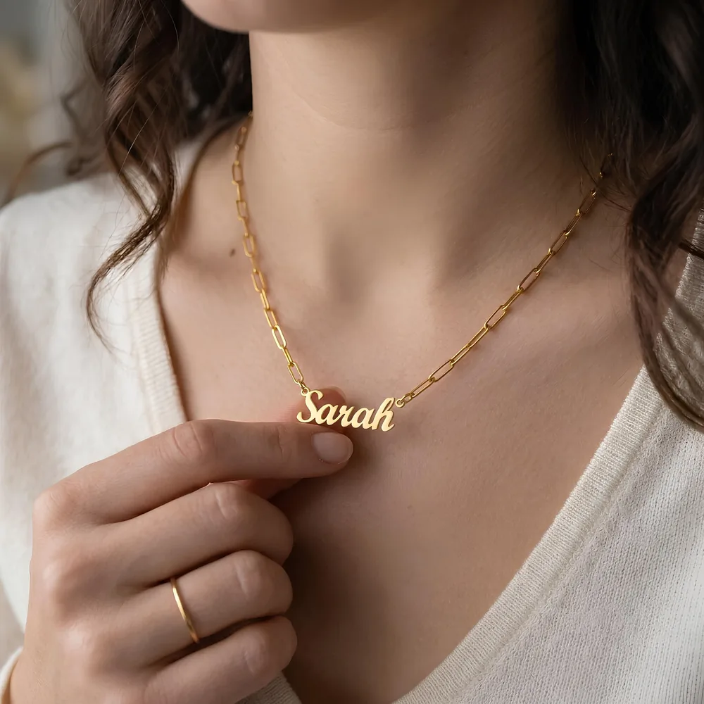 Woman in a cream cardigan holding a gold name pendant that reads Sarah on a paperclip-chain necklace