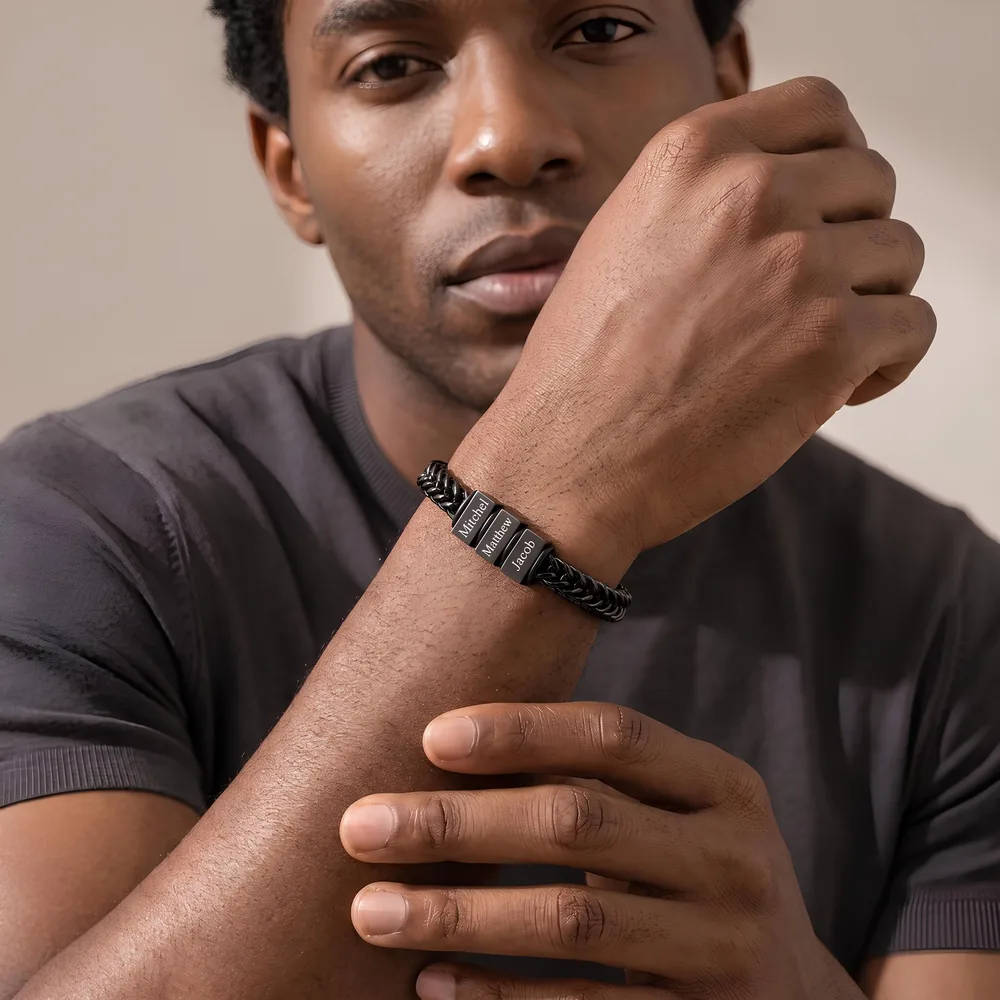A man resting his wrist near his face, wearing a black braided bracelet engraved with three names