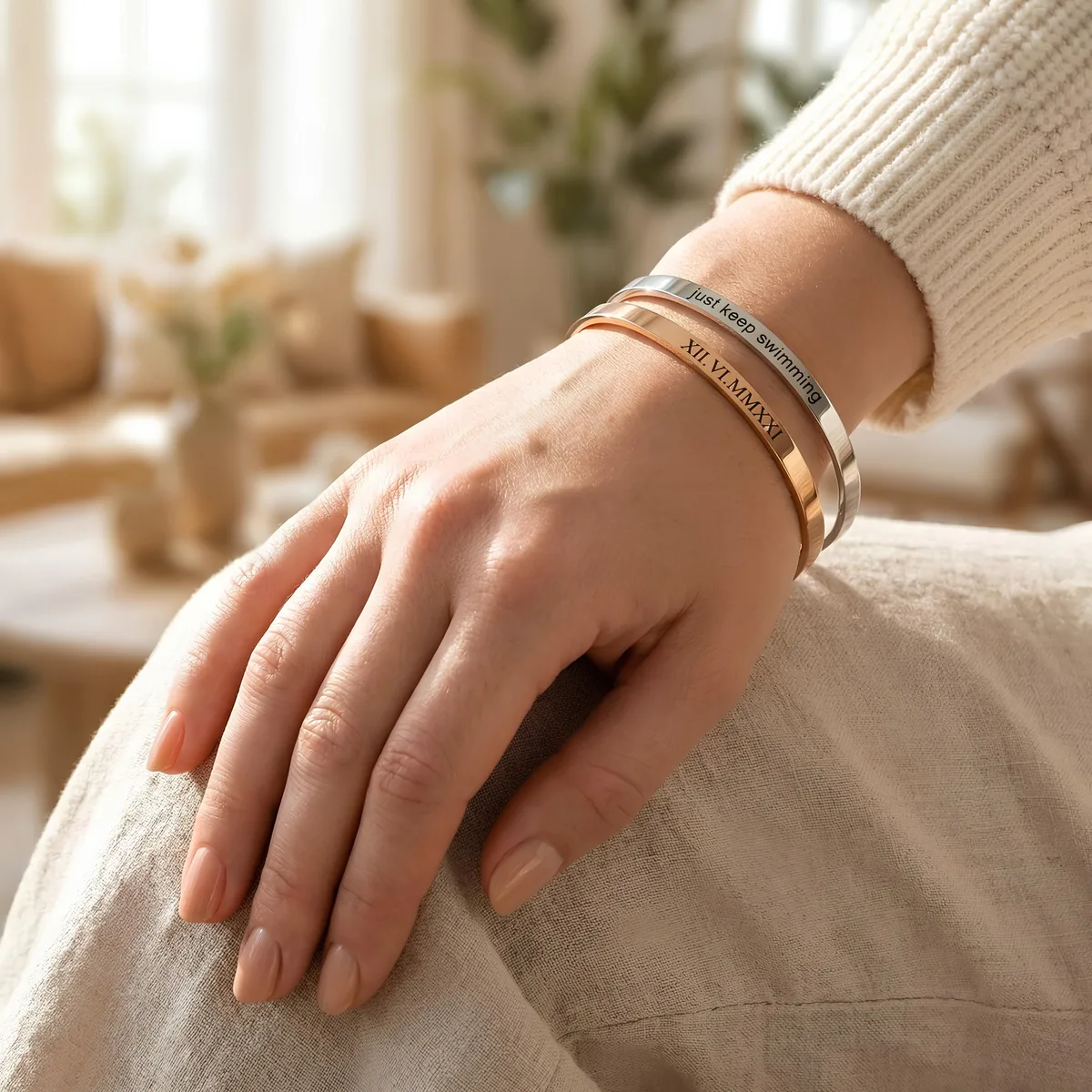 Hand resting on a knee wearing two stacked engraved cuff bracelets, one silver and one rose gold, in a cozy living room